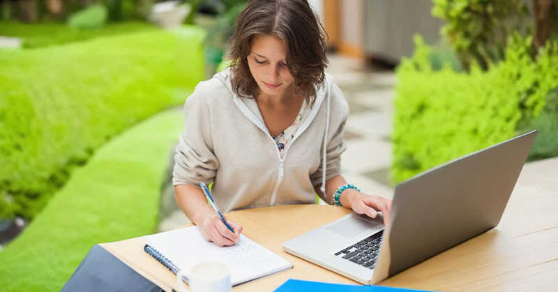 Une jeune femme  travaillant sur un ordinateur portable ou Laptop à l'extérieur en prenant des notes.